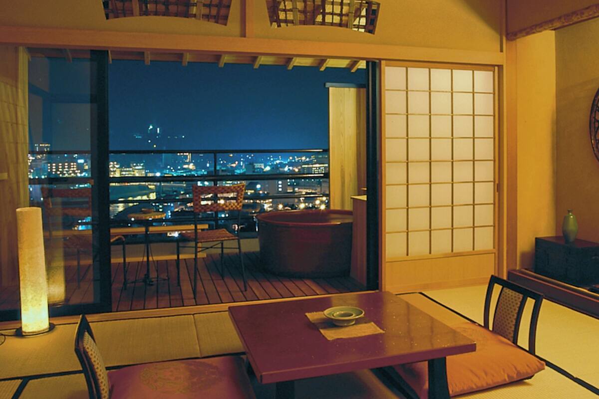 Ein Zimmer mit Blick auf eine beleuchtete Stadtlandschaft bei Nacht, eine Schiebetür, ein Tisch mit Kissen und ein traditioneller japanischer Bildschirm.