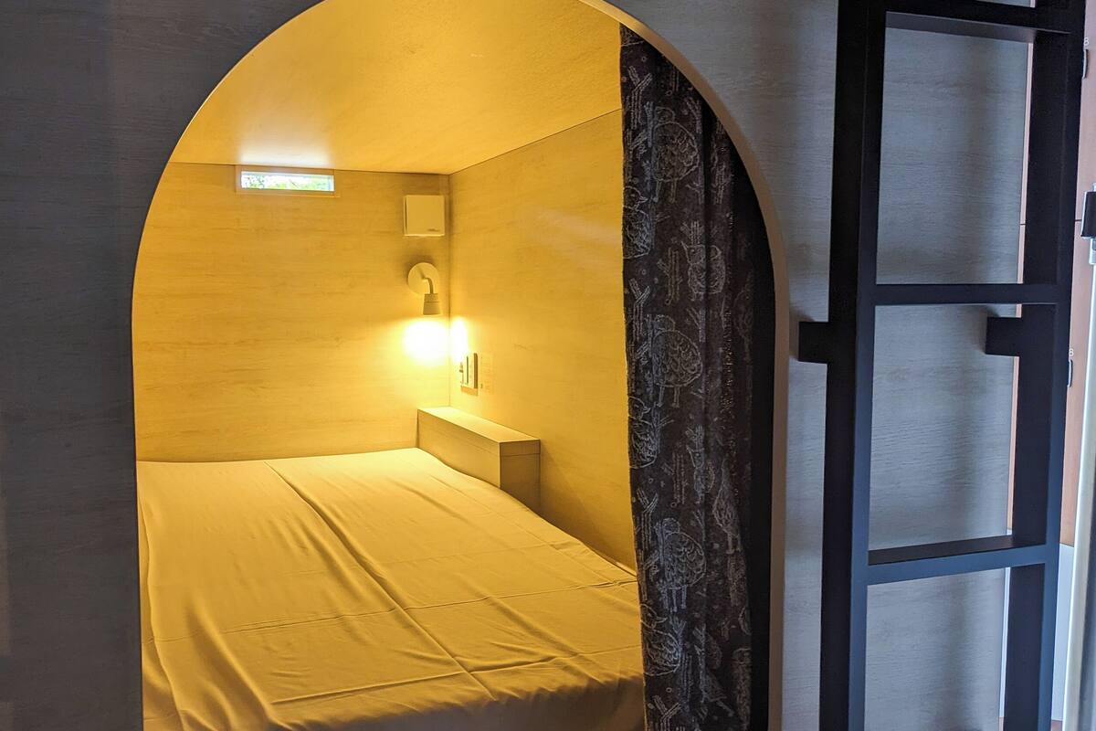 Ein Einzelbett in einem Raum mit gebogener Wand, einem kleinen Fenster und einer wandmontierten Leuchte.