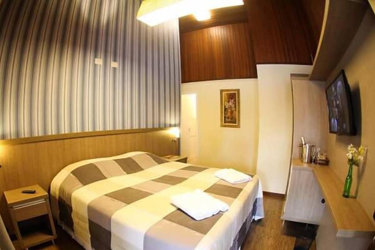 Een hotelkamer met een groot bed, een nachtkastje, een bijzettafeltje, een spiegel, een inlijsting foto aan de muur en een kleintje tafeltje met een bloevas.
