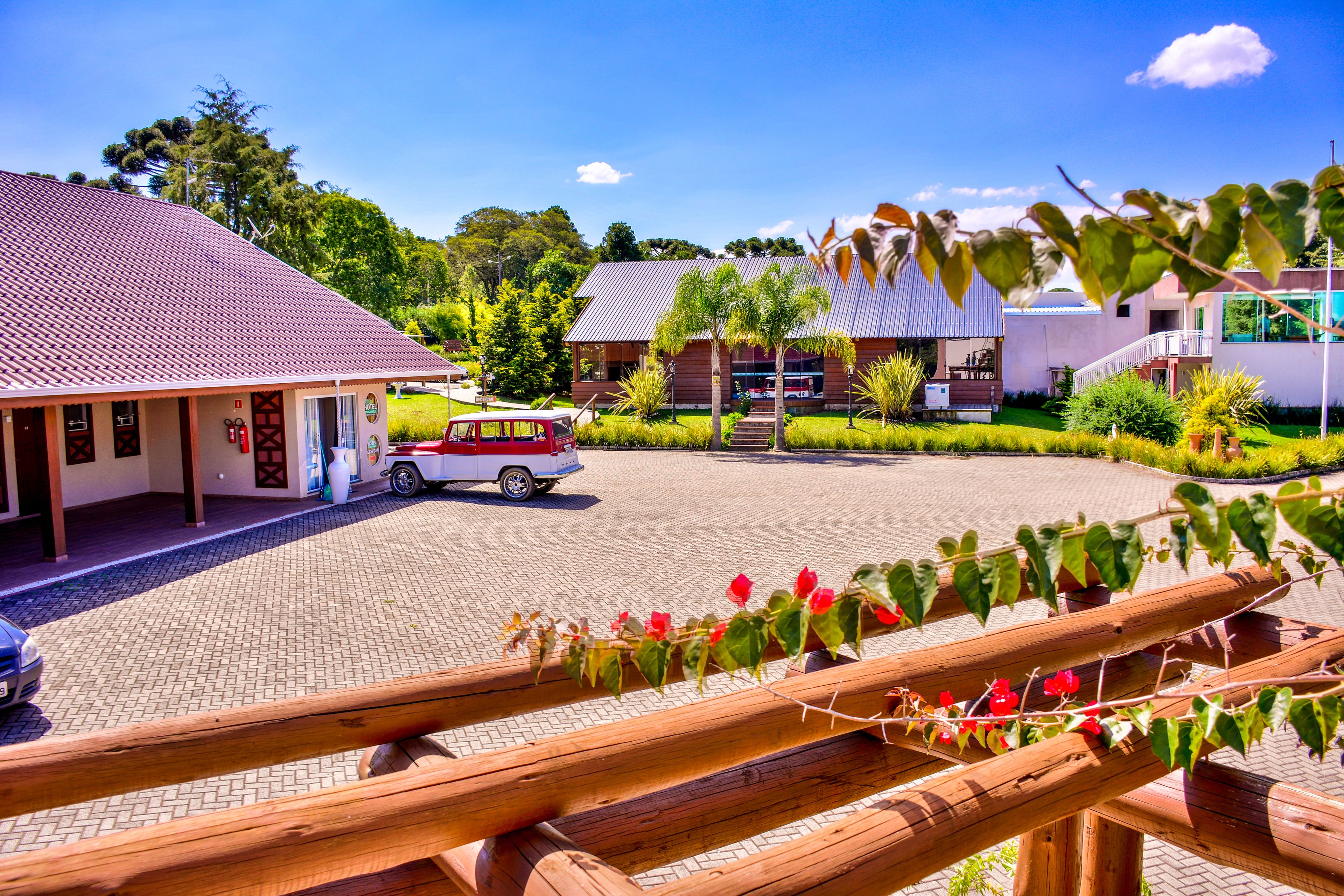 Um hotel com calçada de acesso, um carro vermelho estacionado em frente e uma cerca de madeira com flores vermelhas.