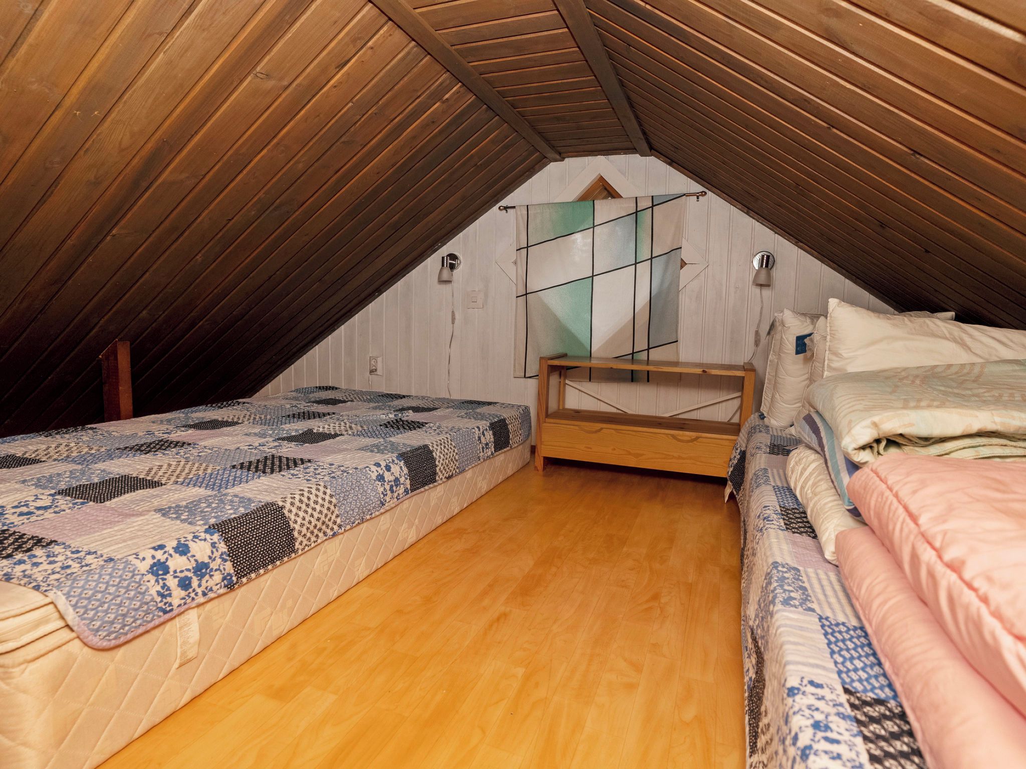 A small attic bedroom with a sloped ceiling, wooden flooring, a bed with a patterned quilt, a bedside table, and a wall-mounted shelf.