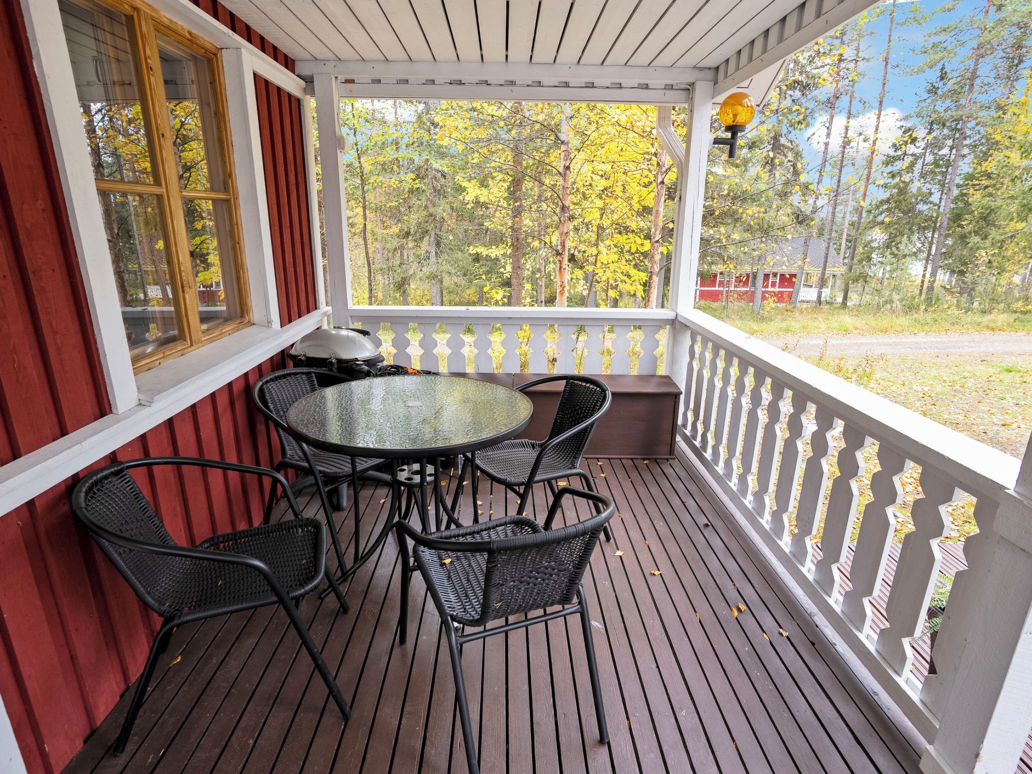 A covered outdoor seating area with a table and chairs, a view of trees, and a red building in the background.