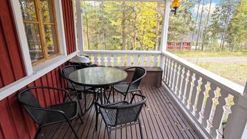 A covered outdoor seating area with a table and chairs, a view of trees, and a red building in the background.