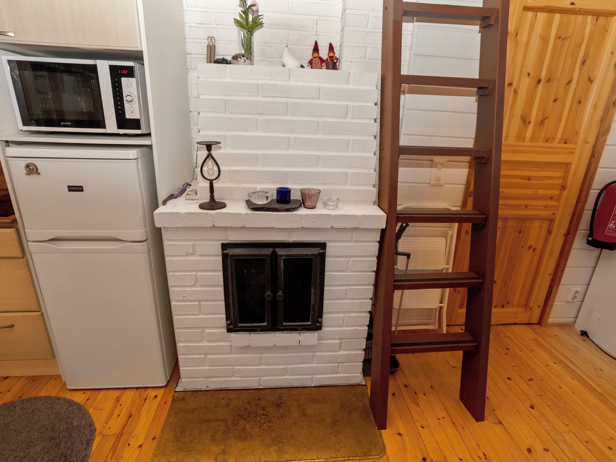 Cocina compacta con chimenea de ladrillo blanco, microondas y refrigerador. Una escalera de madera está apoyada contra la pared, y hay un estante con objetos decorativos sobre la chimenea.