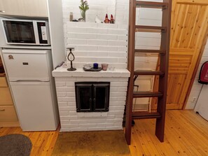 Cocina compacta con chimenea de ladrillo blanco, microondas y refrigerador. Una escalera de madera está apoyada contra la pared, y hay un estante con objetos decorativos sobre la chimenea.