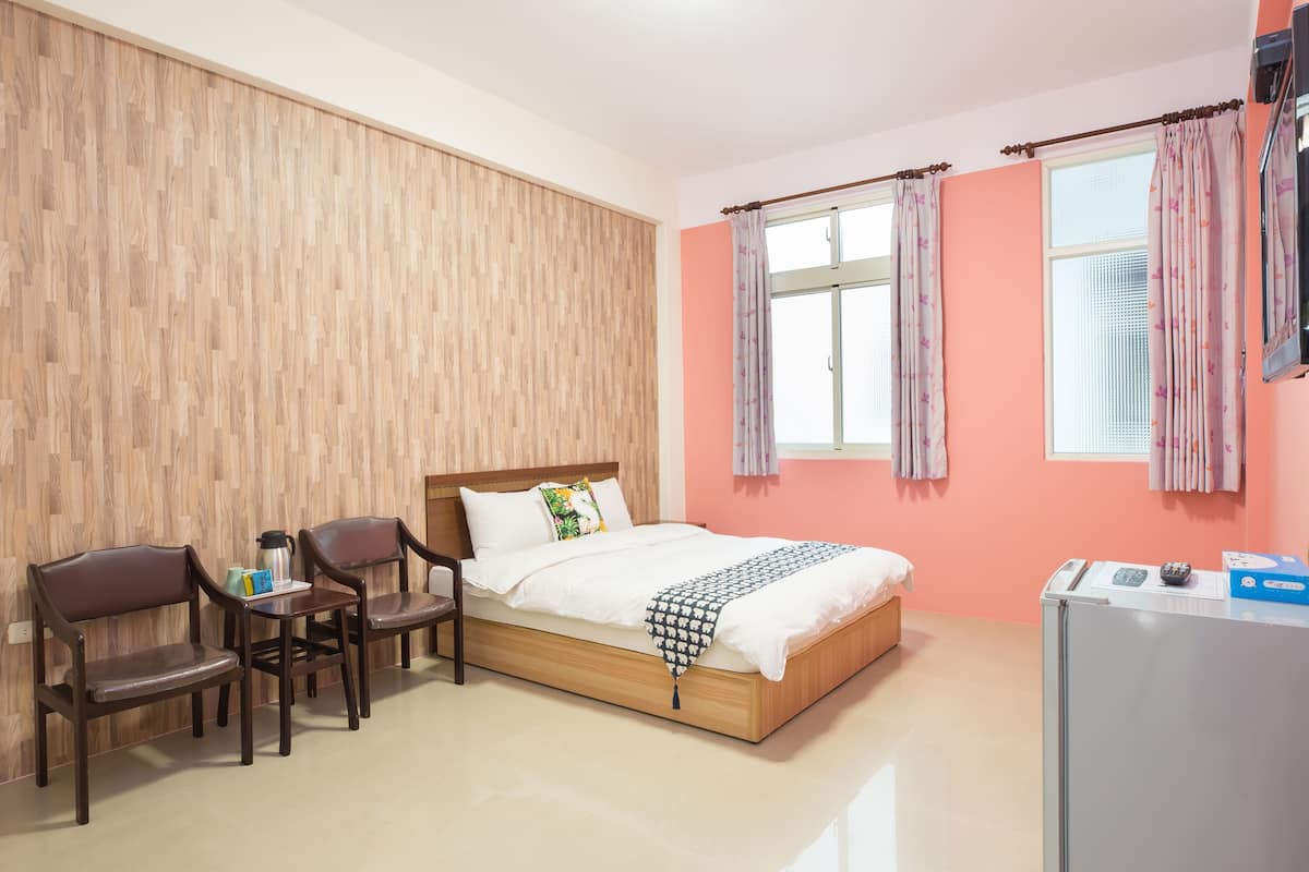 A bedroom with a wooden bed, two chairs, a small table with a kettle, and a refrigerator.