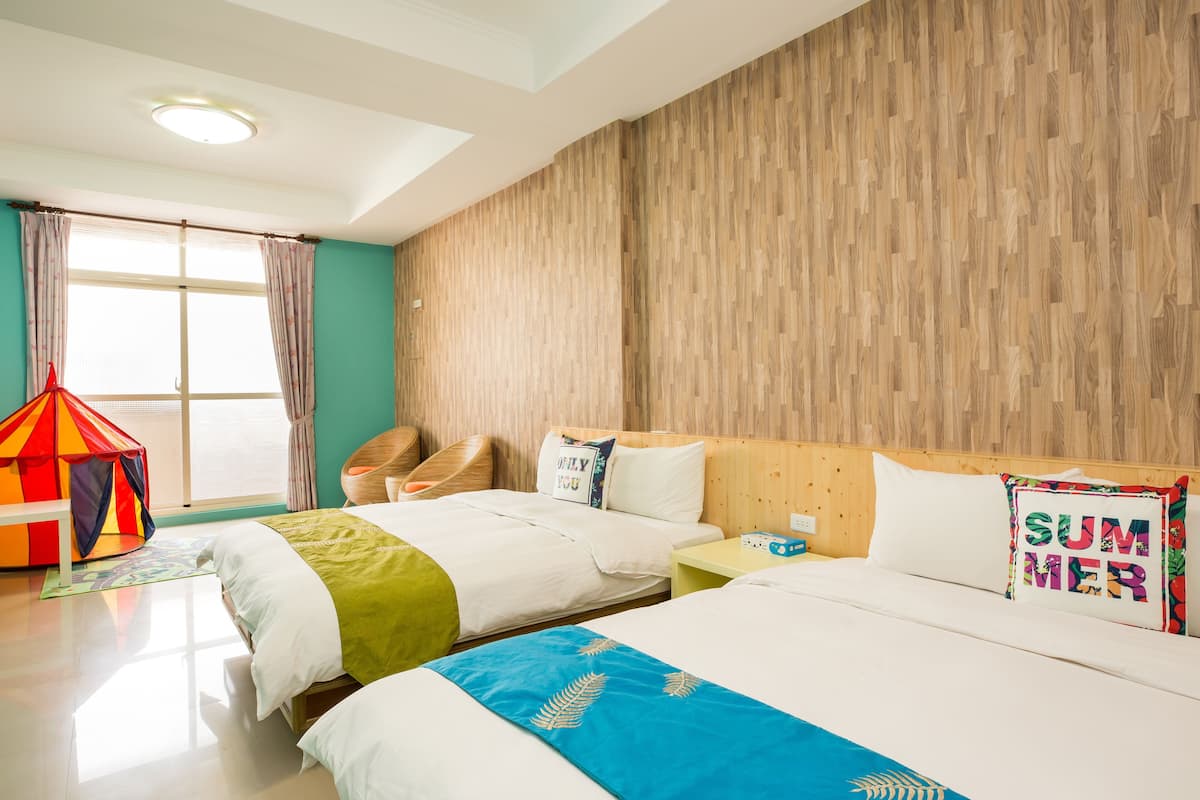A hotel room with two beds, a wooden wall, a window with curtains, a small table, and a colorful cushion.