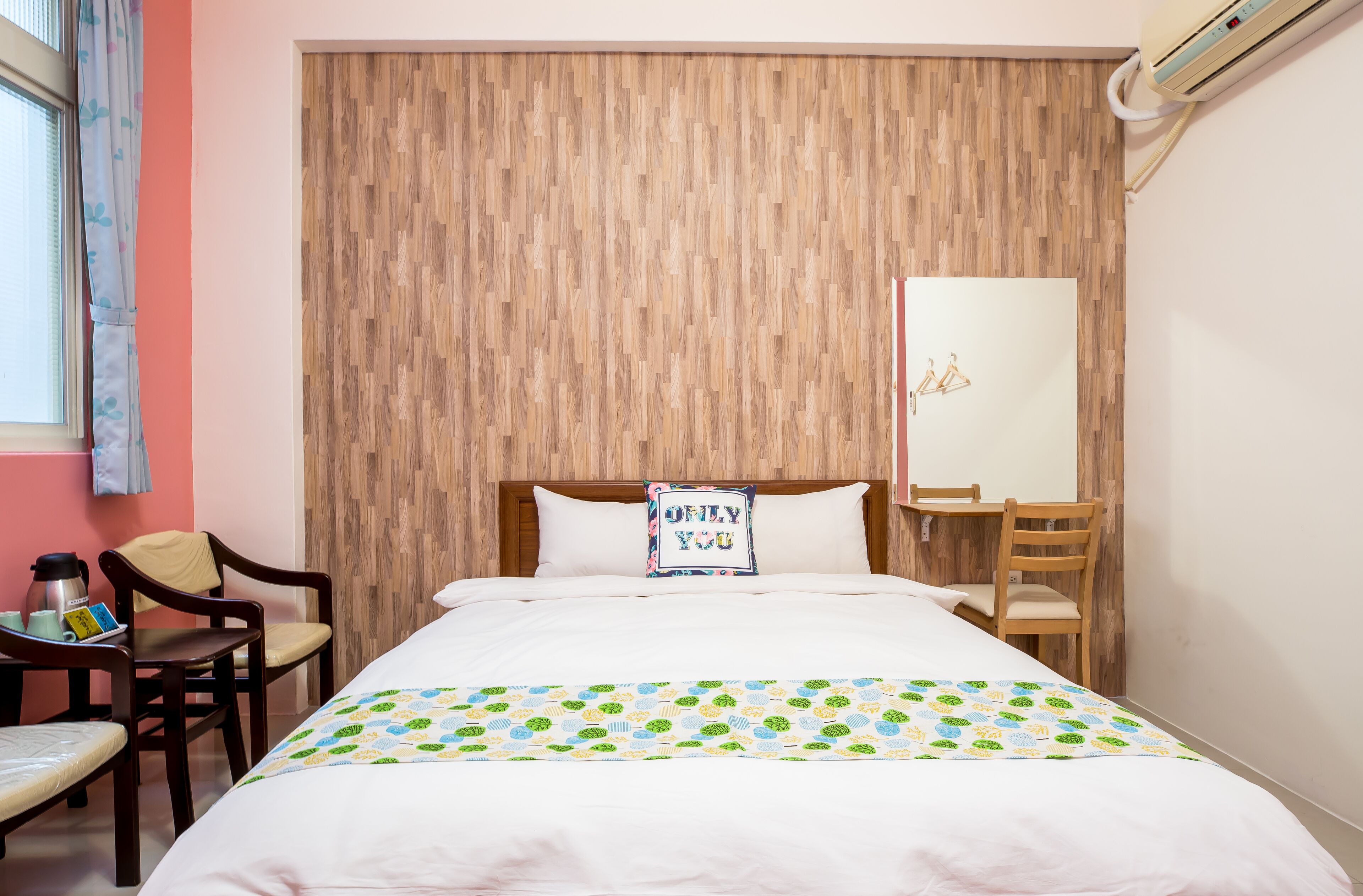 A hotel room with a wooden headboard, a bed with a white comforter, a bedside table with a coffee maker, and a chair.