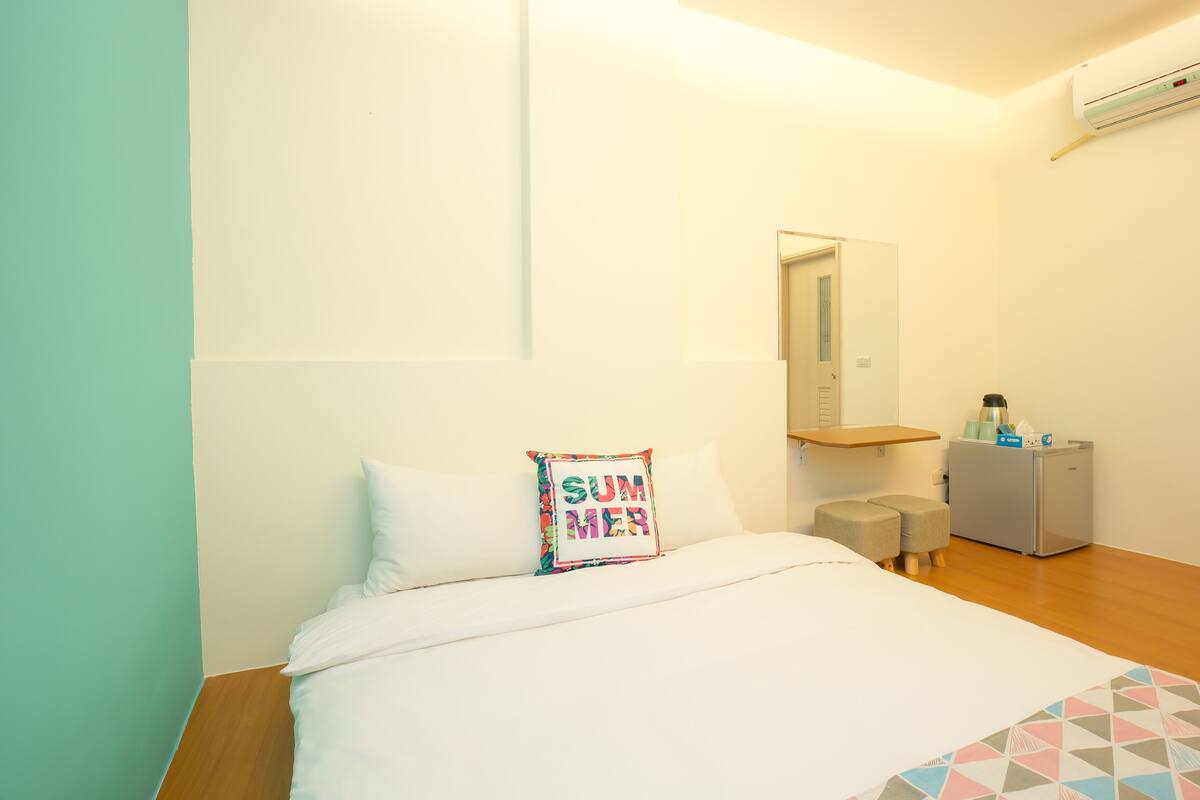 A bedroom with a bed, a small desk, a mirror, and a refrigerator.