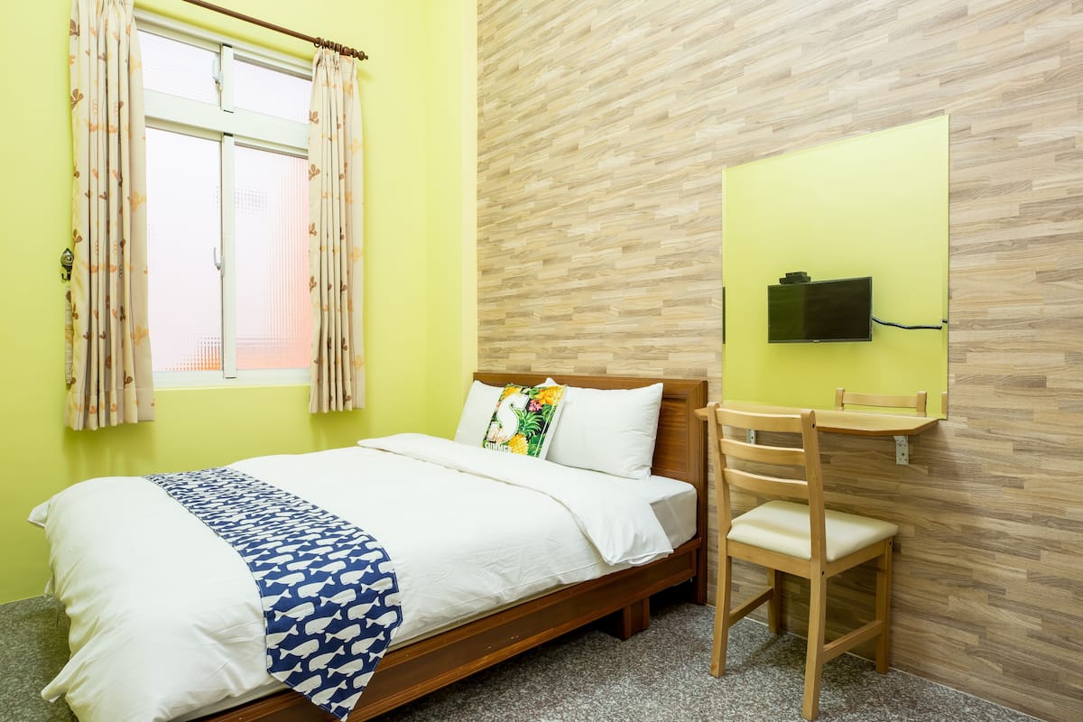 A room with a bed, a wooden chair, a television mounted on the wall, and a window with curtains.