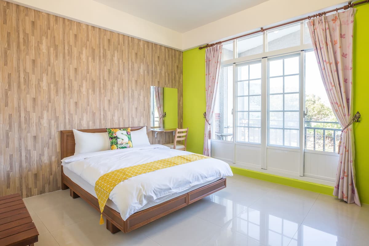 A bedroom with a wooden headboard, a bed with white bedding and a yellow throw, a wooden nightstand, a chair, and a large window with curtains.