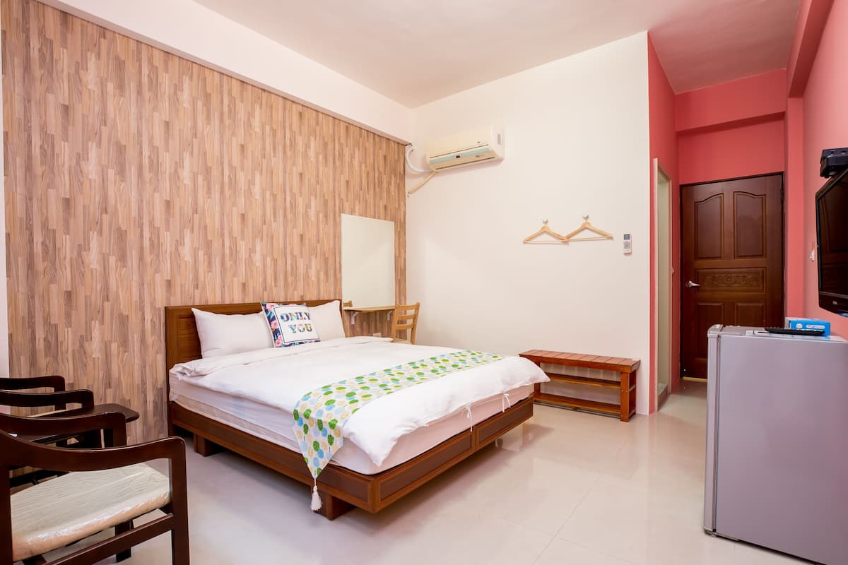 A bedroom with a wooden headboard, a bed with a white comforter, a small bedside table, a bench, and a refrigerator.