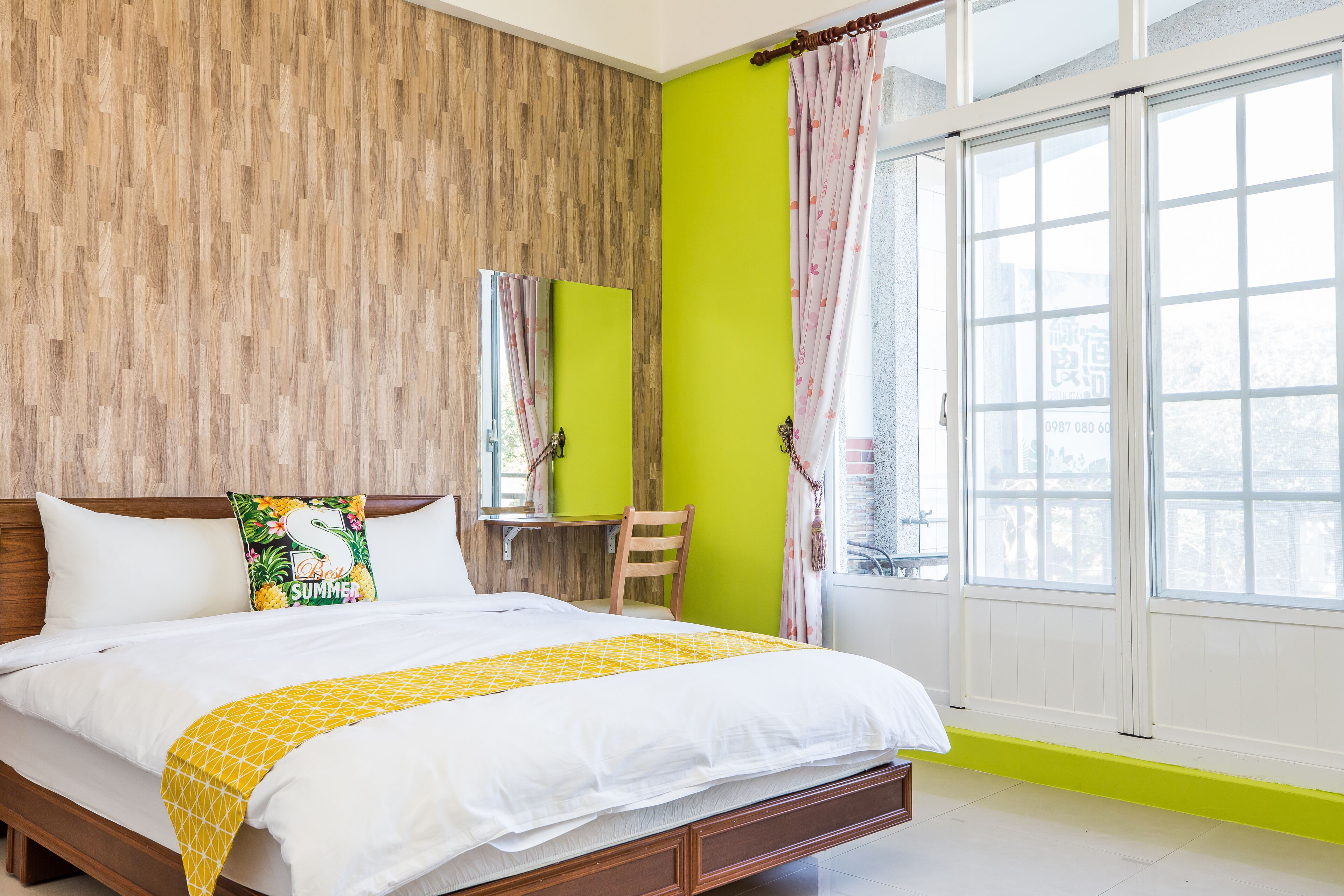 A bedroom with a wooden headboard, a bed with white bedding and a yellow accent, a small desk with a chair, and a large window with curtains.