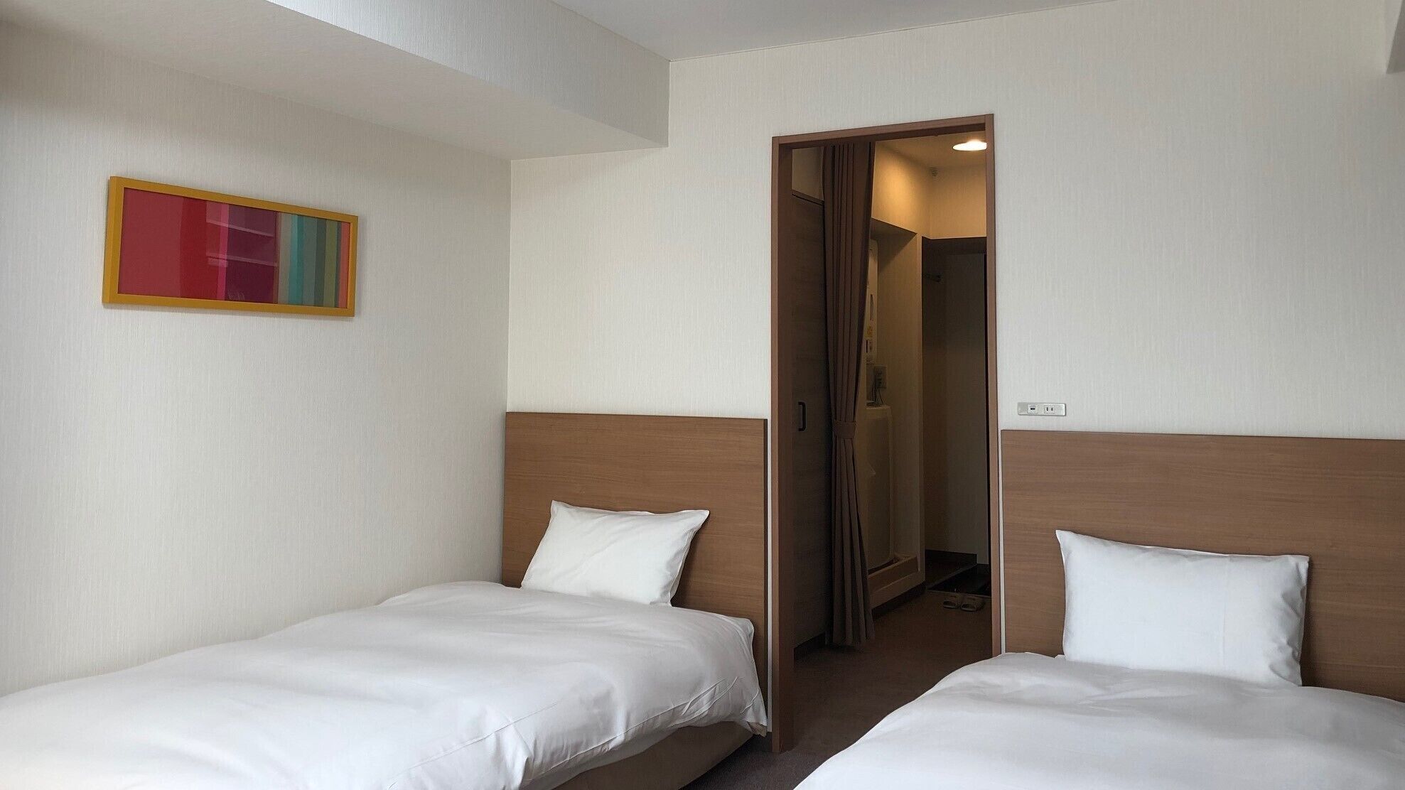 A hotel room with two single beds, a door leading to a bathroom, and a colorful painting on the wall.
