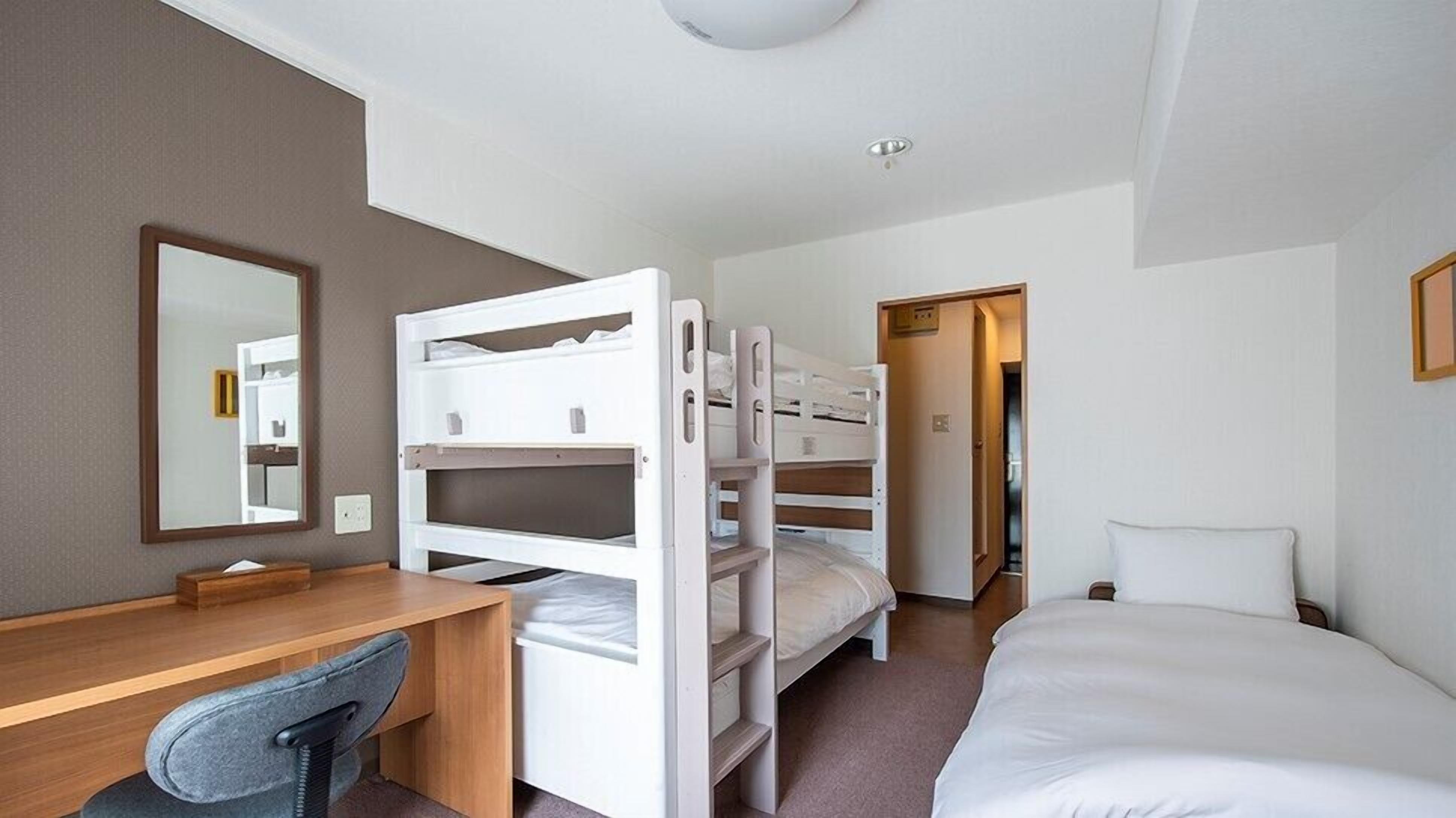 A hotel room with bunk beds, a desk, a chair, a bed, a mirror, and a door leading to another room.