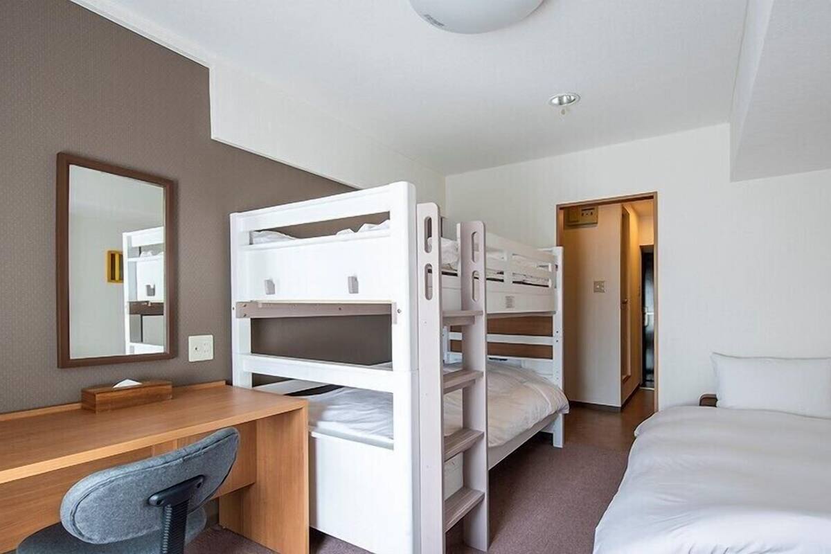 A hotel room with bunk beds, a desk, a chair, a bed, a mirror, and a door leading to another room.