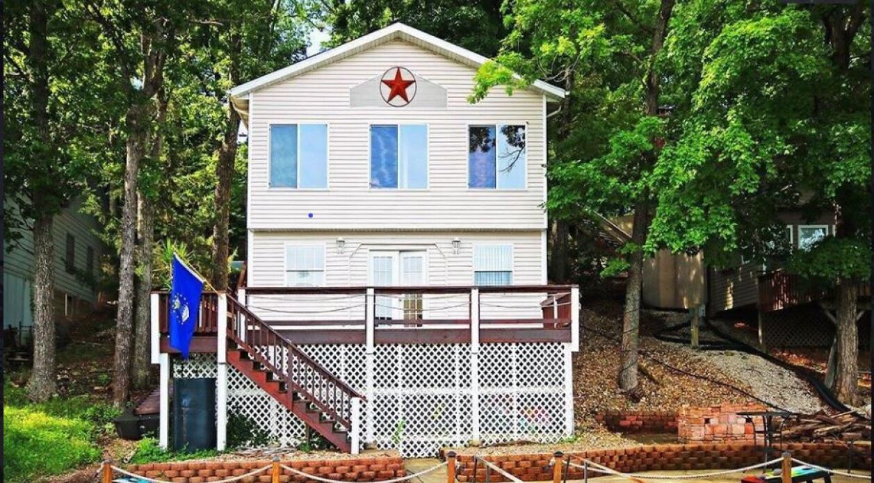 Une maison à deux étages, avec une façade blanche, une étoile rouge sur le toit et une terrasse en bois entourée d’une clôture blanche.