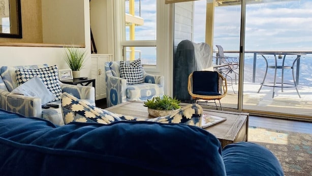 Een woonkamer met een grote bank, decoratieve kussens, een bijzettafeltje met een plant, en uitzicht op de natuur via een schuifdeur.