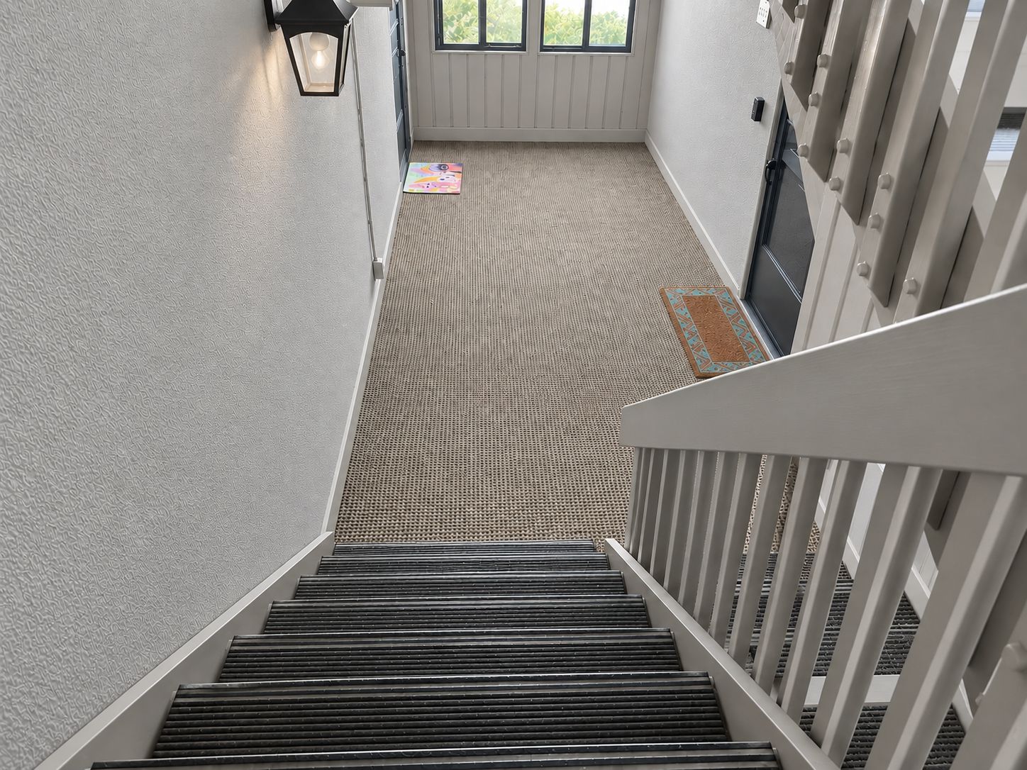 A staircase leading down to a carpeted area with a doormat and a wall-mounted light fixture.