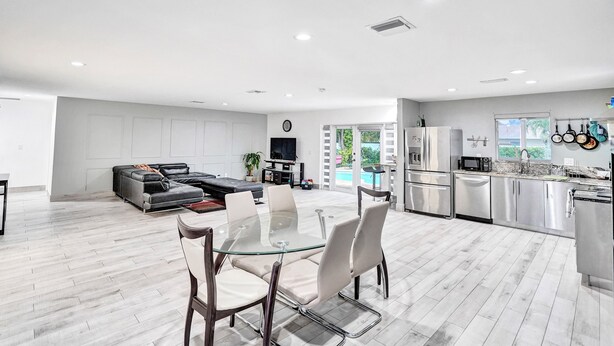 A modern kitchen with stainless steel appliances, a dining area with a glass table, and a living room with a sofa and TV.