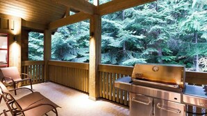 Ein überdachter Außenbereich mit Grill, Stühlen und Blick auf dichten Wald.