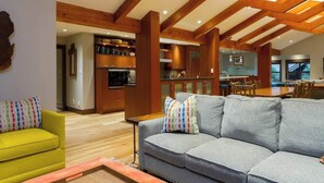 Ein modernes Interieur mit Holzbalkendecke, einem grauen Sofa mit bunten Kissen, einem gelben Sessel und einer Küchenzeile mit Holzausstattung sowie einem Esstisch.