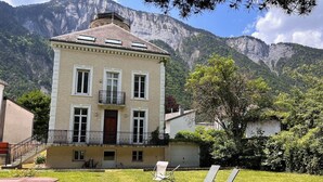 A two-story building with a balcony, a small garden, and a mountain backdrop. - STUNNING FRENCH HOUSE, SPACIOUS AND WELL EQUIPPED - SLEEPS 10 (Le Bourg-d'Oisans)