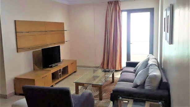 A living room with a sofa set, a TV on a wooden stand, a glass coffee table, and a large window with curtains. - Sea view Serviced Apartment 2 bedroom 2 bath #1 (Hawally)
