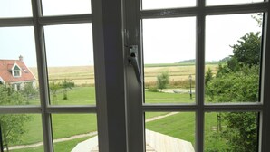 Blick durch ein Fenster auf eine Rasenfläche, eine Holzterrasse und ein entfernteres Gebäude mit rotem Dach.