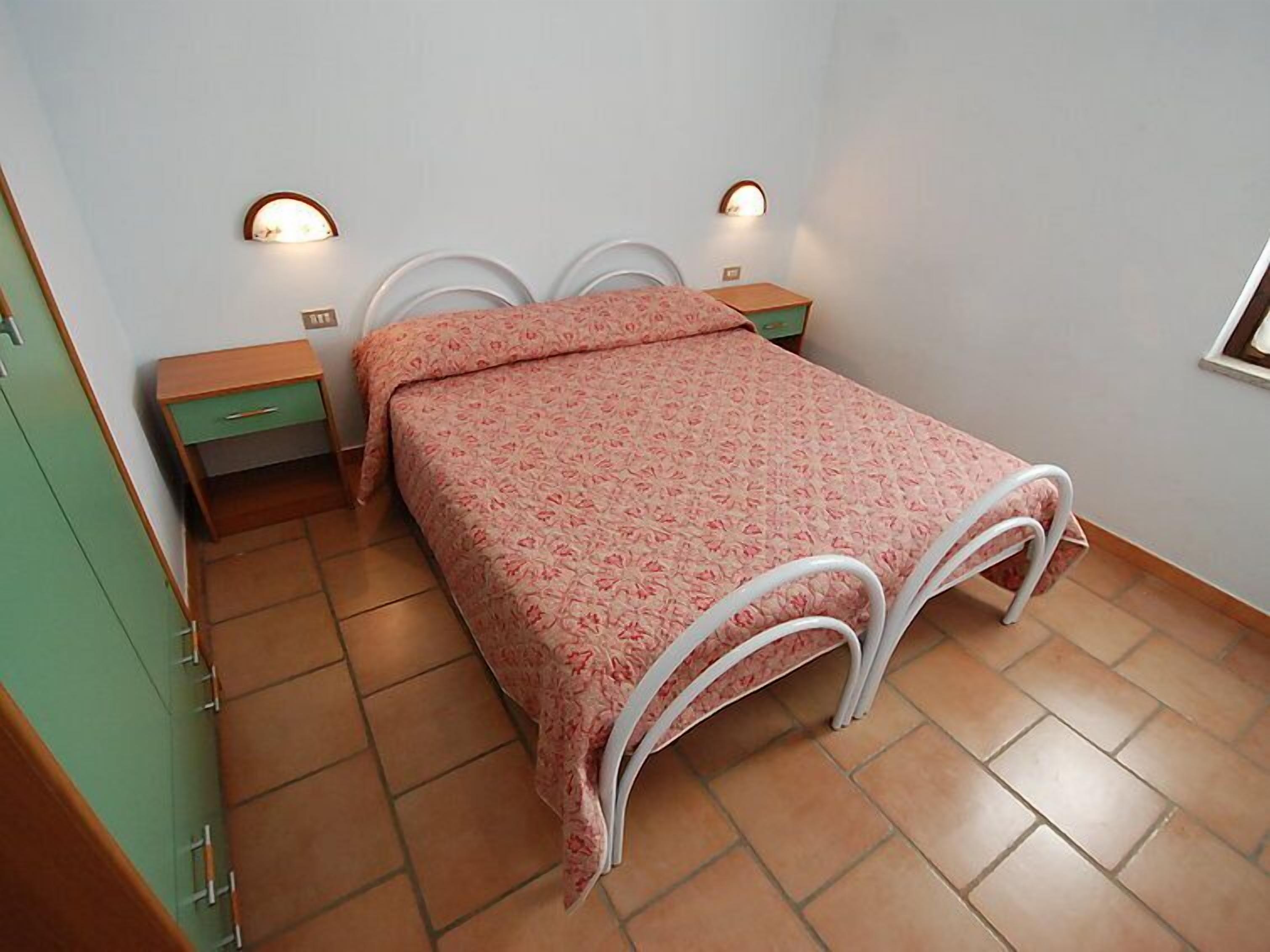 A single-bed room with a patterned bedspread, two bedside tables, a green wardrobe, and two wall-mounted lights.