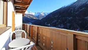 A balcony with wooden railings and a white plastic chair, overlooking a mountainous landscape with snow-capped peaks.