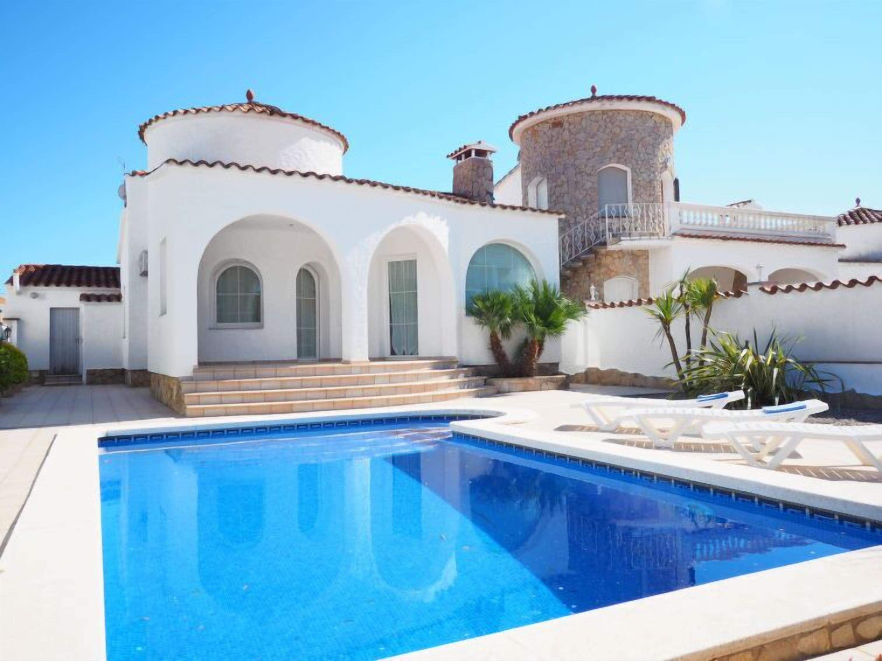 Un espace piscine avec une eau d’un bleu limpide, un bâtiment blanc doté de fenêtres en arc de cercle et une tour en pierre.