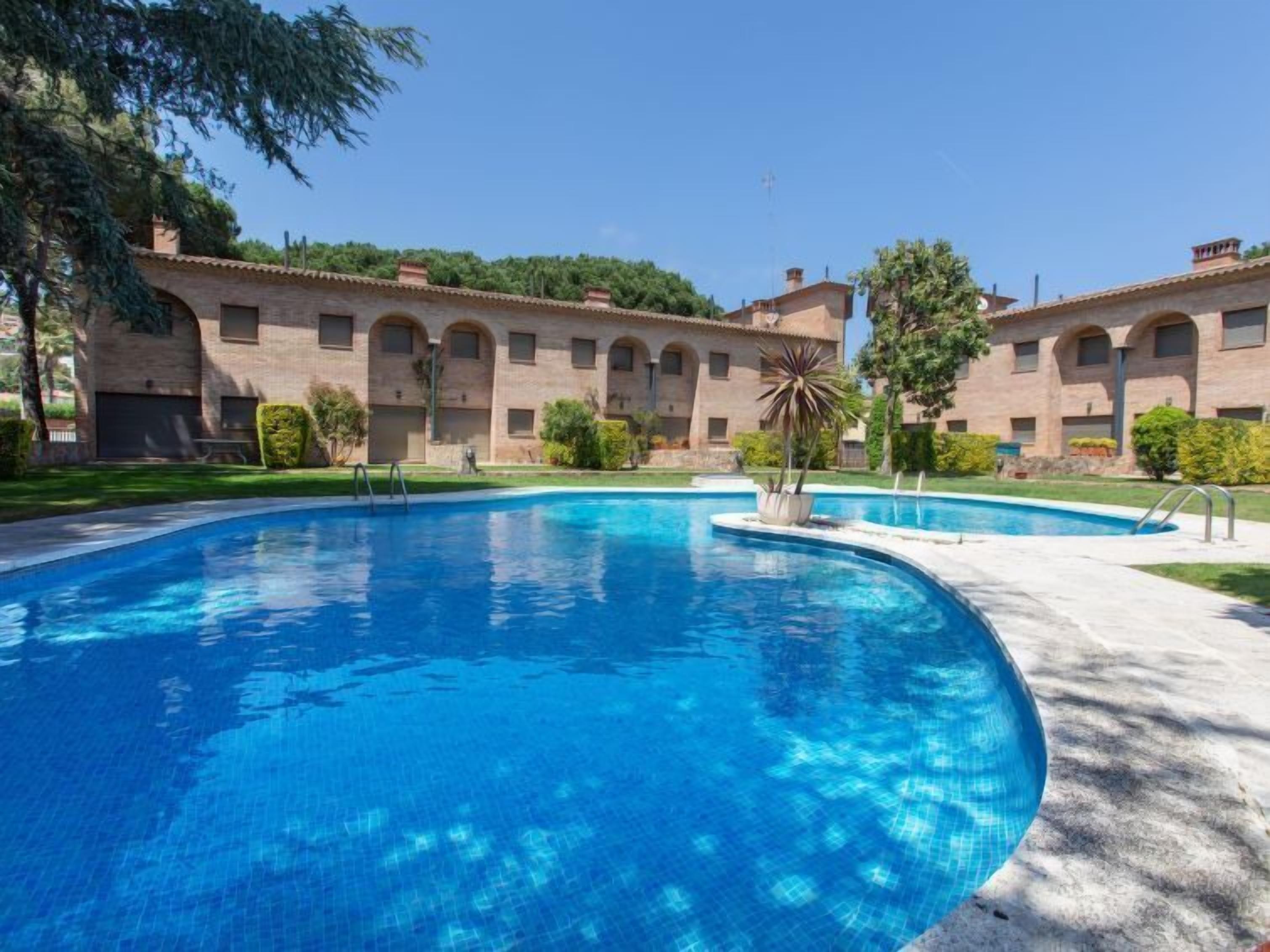 A swimming pool with a curved edge, surrounded by a paved area. There are two buildings with arched windows and a brick facade in the background.