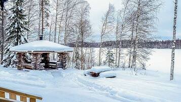 Lumihunnun peittämä hirsimökki, jonka ympärillä on lumipeitteinen maisema ja puita.