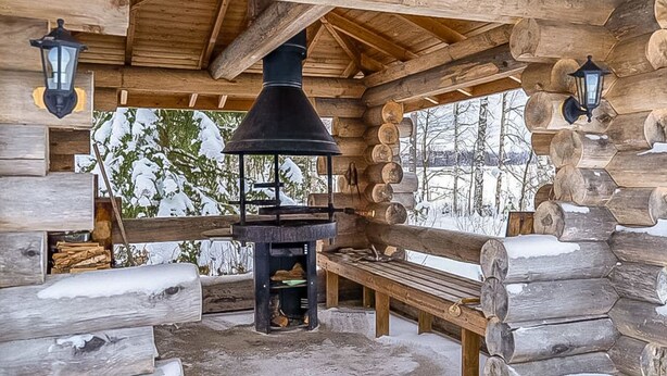 Una baita di legno con un camino al centro, panche e due lanterne fissate alle pareti.