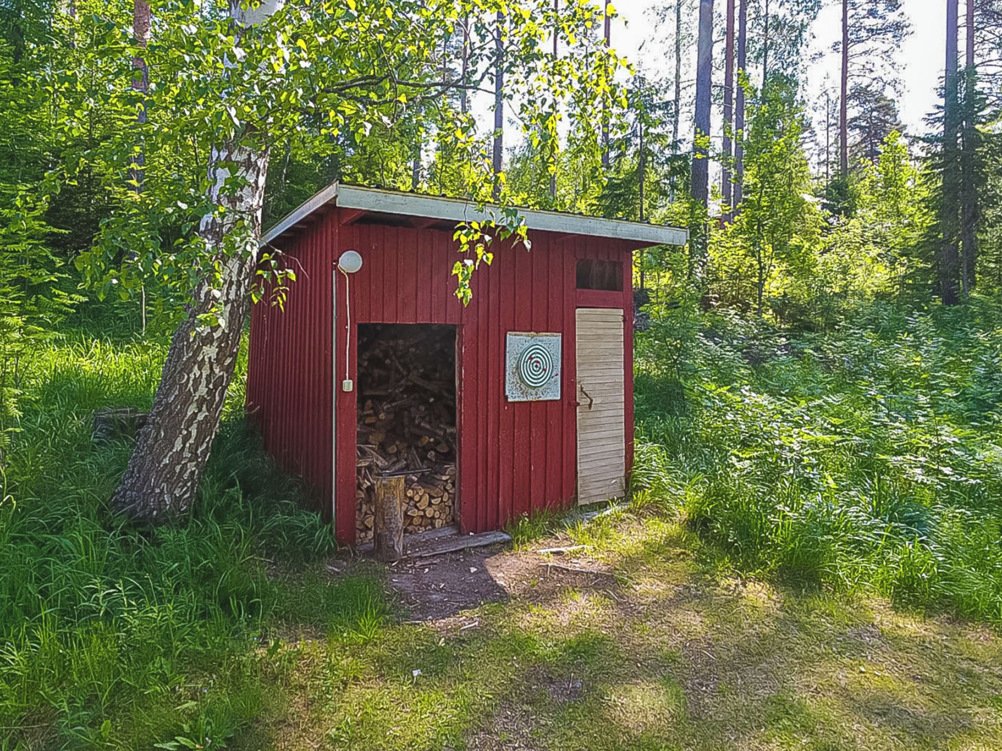 A small red wooden shed with a door and a window, surrounded by greenery and trees.