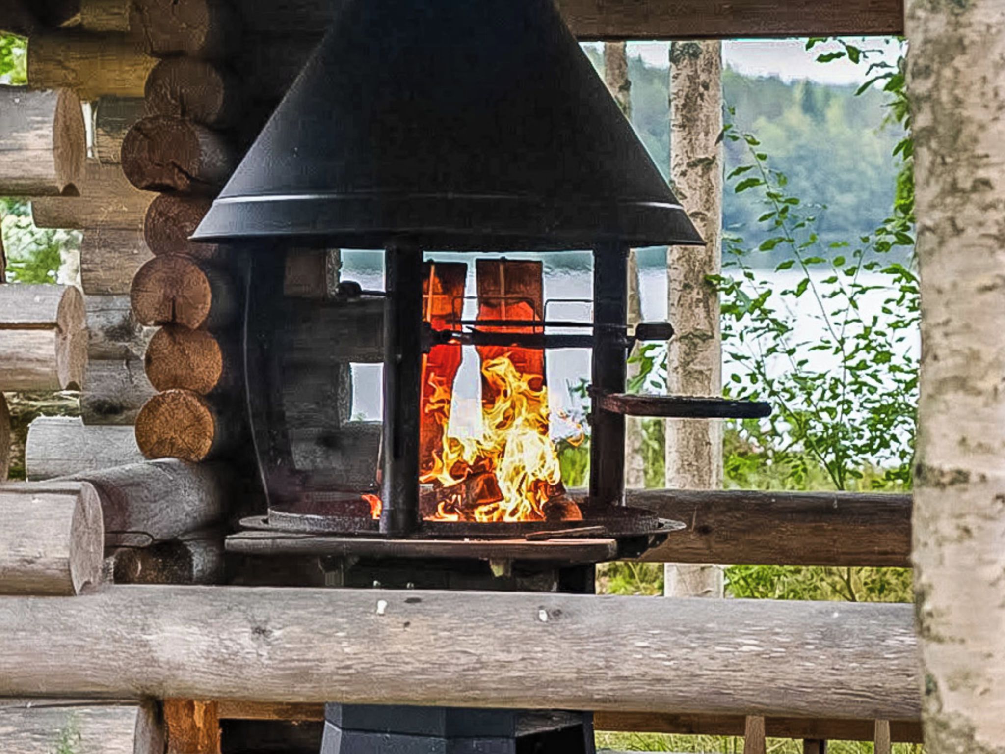 A wood-burning stove with logs stacked to the left, set in a wooden structure with a view of greenery in the background.