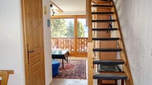 Una escalera de madera lleva a un balcón con una mesa y sillas, un tapete estampado y vistas a árboles a través de un ventanal.