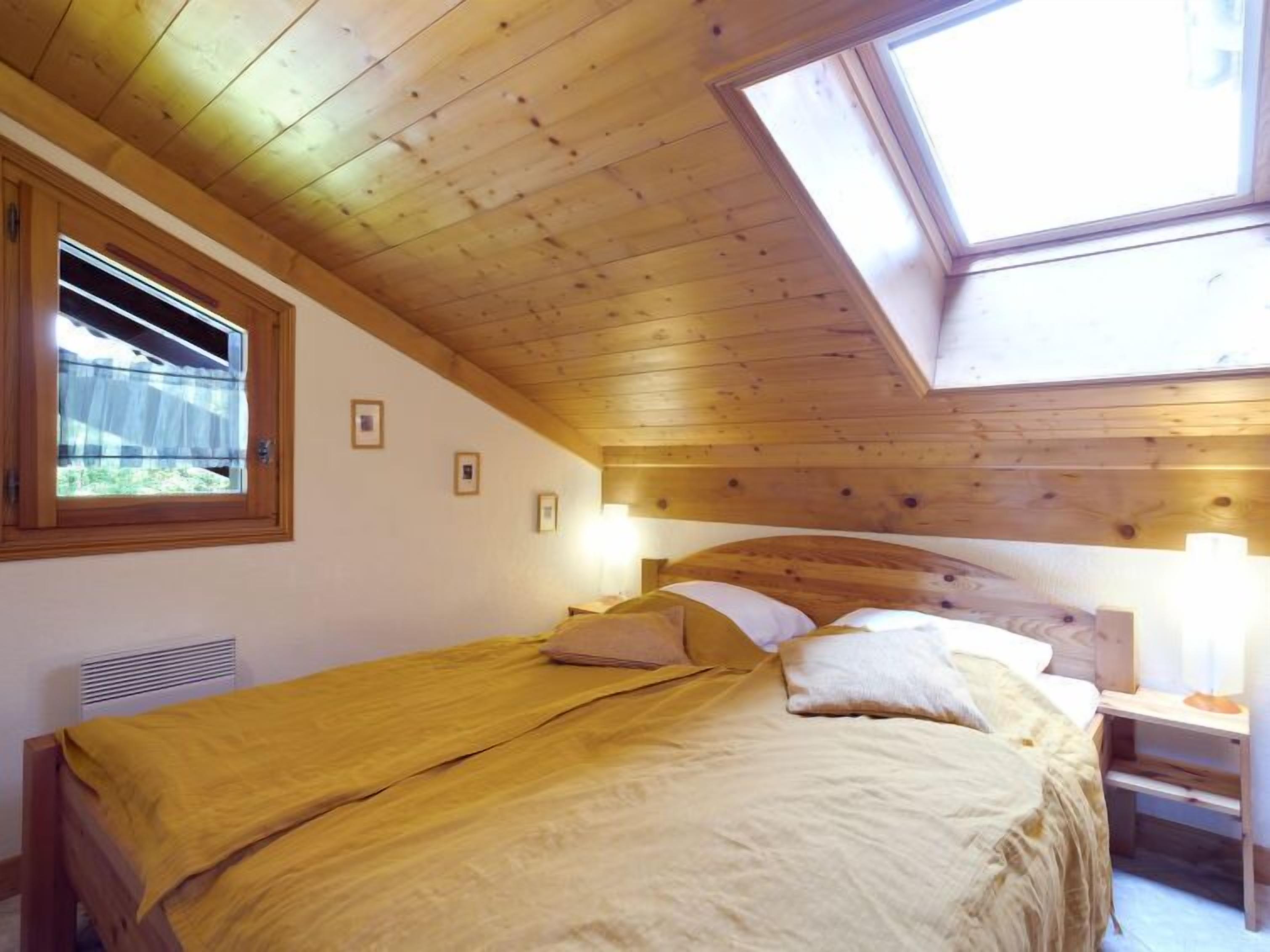 A bedroom with a sloped ceiling, wooden beams, a bed with a yellow bedspread, a window with blinds, and a bedside table with a lamp.