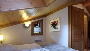 Un dormitorio con techo inclinado, cama con ropa de cama blanca, una obra de arte enmarco, una ventana y una puerta de madera.