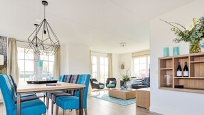 A modern living room with a dining area, featuring a wooden dining table, blue upholstered chairs, a geometric pendant light, and a wooden coffee table. - Tapuit 6 by Interhome (Noordwijk)