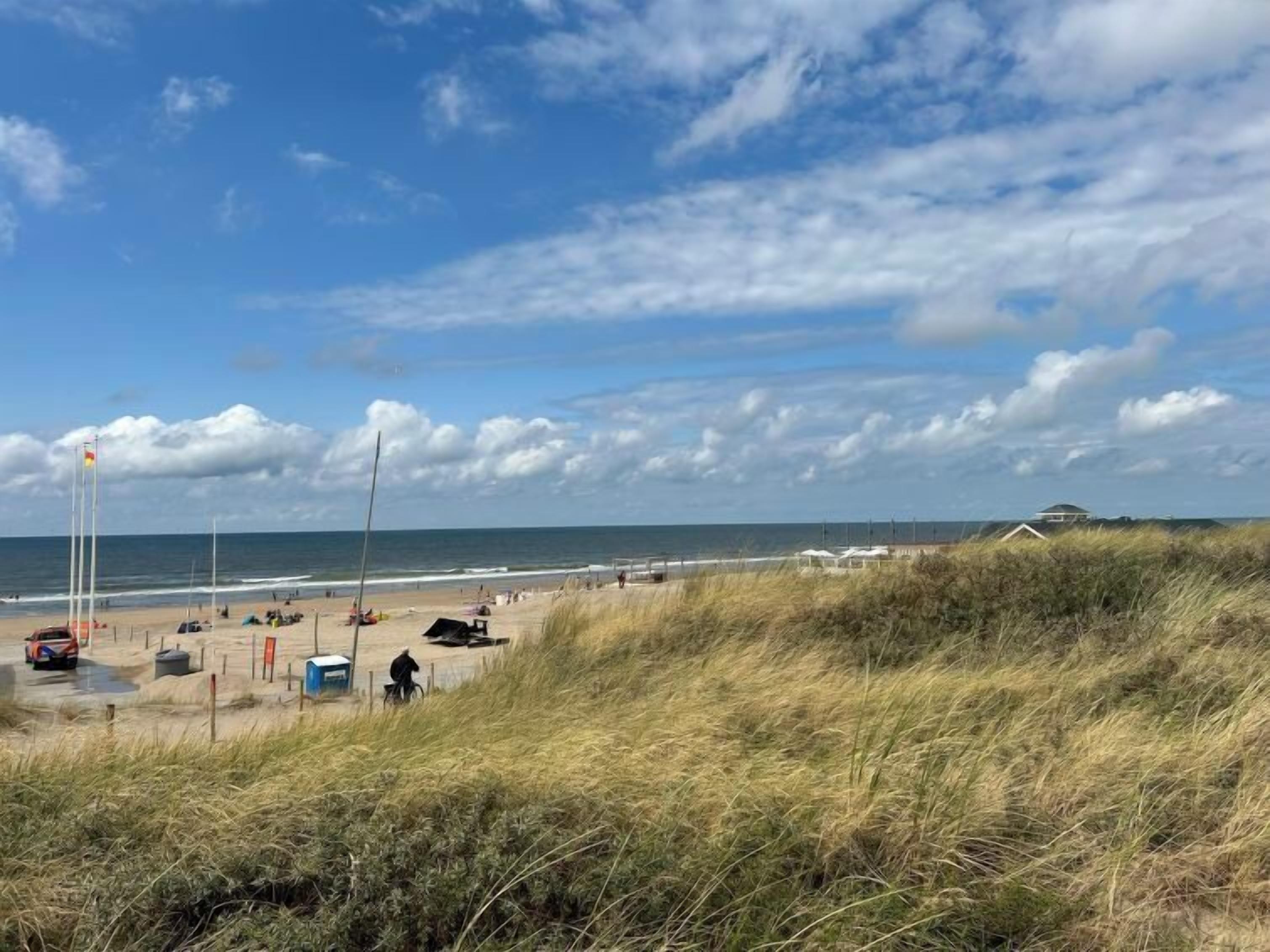 Een strand met duinen, een vlaggenmast en een huis in de verte.