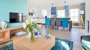 A modern living room with a flat-screen TV, a wooden coffee table with a vase of flowers, and a dining area with blue chairs. - Tapuit 6 by Interhome (Noordwijk)