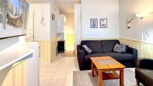 Sala de estar com sofá escuro, mesa de centro de madeira e televisão montada na parede. Há uma porta que dá para outro cômodo e um abajur no lado direito.