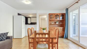 Een moderne woonkamer met een eettafel en stoelen, een grijze bank en een keuken met kastjes en een koelkast.