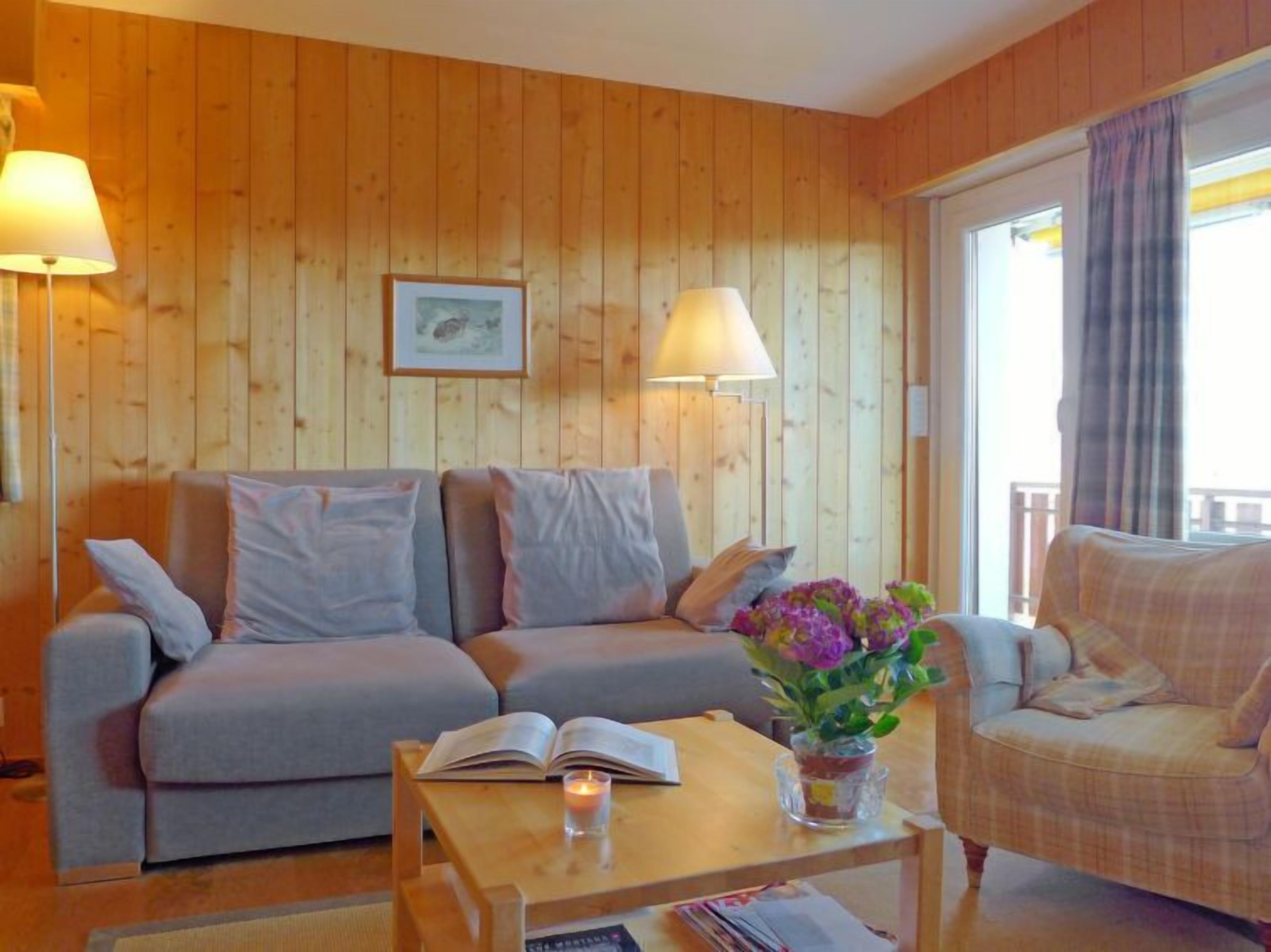 A cozy room with a wooden paneled wall, a grey sofa, a small table with a book and a candle, and a chair with a vase of flowers.