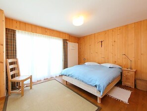 A wooden-paneled bedroom with a bed, a chair, a nightstand, and a window with curtains. - Andrea A 10 by Interhome (Crans-Montana)
