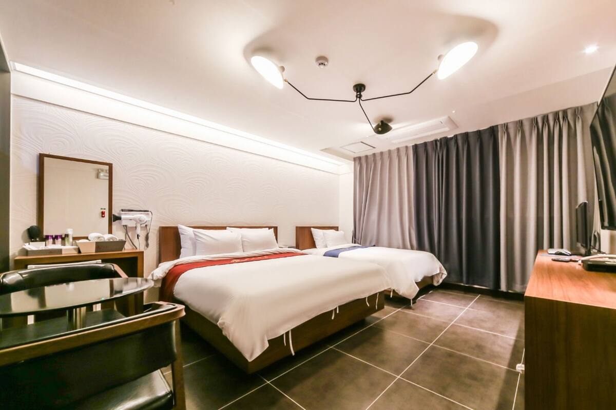 A hotel room with two beds, a desk, a chair, a TV, and a bathroom with a mirror and towel rack.