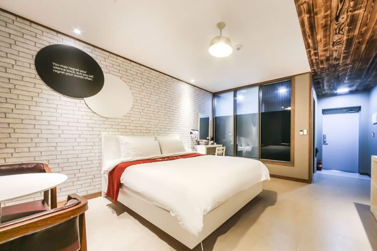 A modern hotel room with a brick wall, a large bed, a desk, and a sliding glass door leading to a balcony.