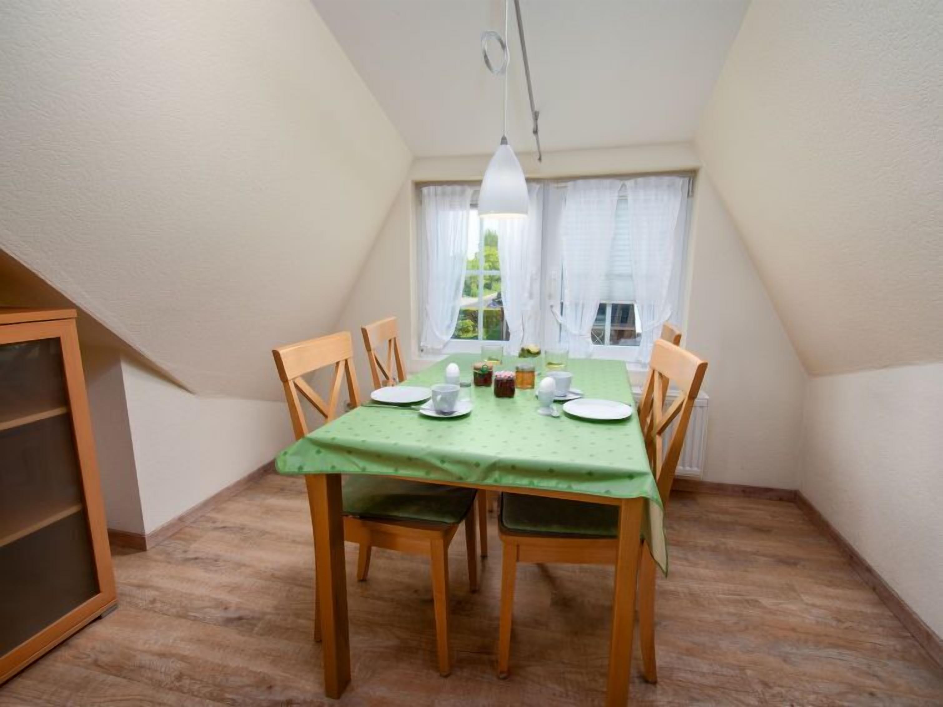 Un coin repas avec une table dressée pour quatre personnes, des chaises en bois, une nappe verte et un luminaire suspendu.